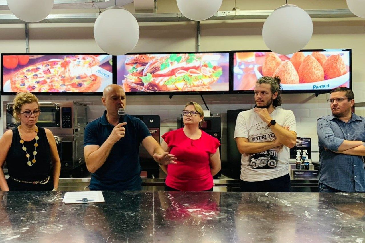 Um grupo de pessoas está em pé ao redor de uma mesa na cozinha.