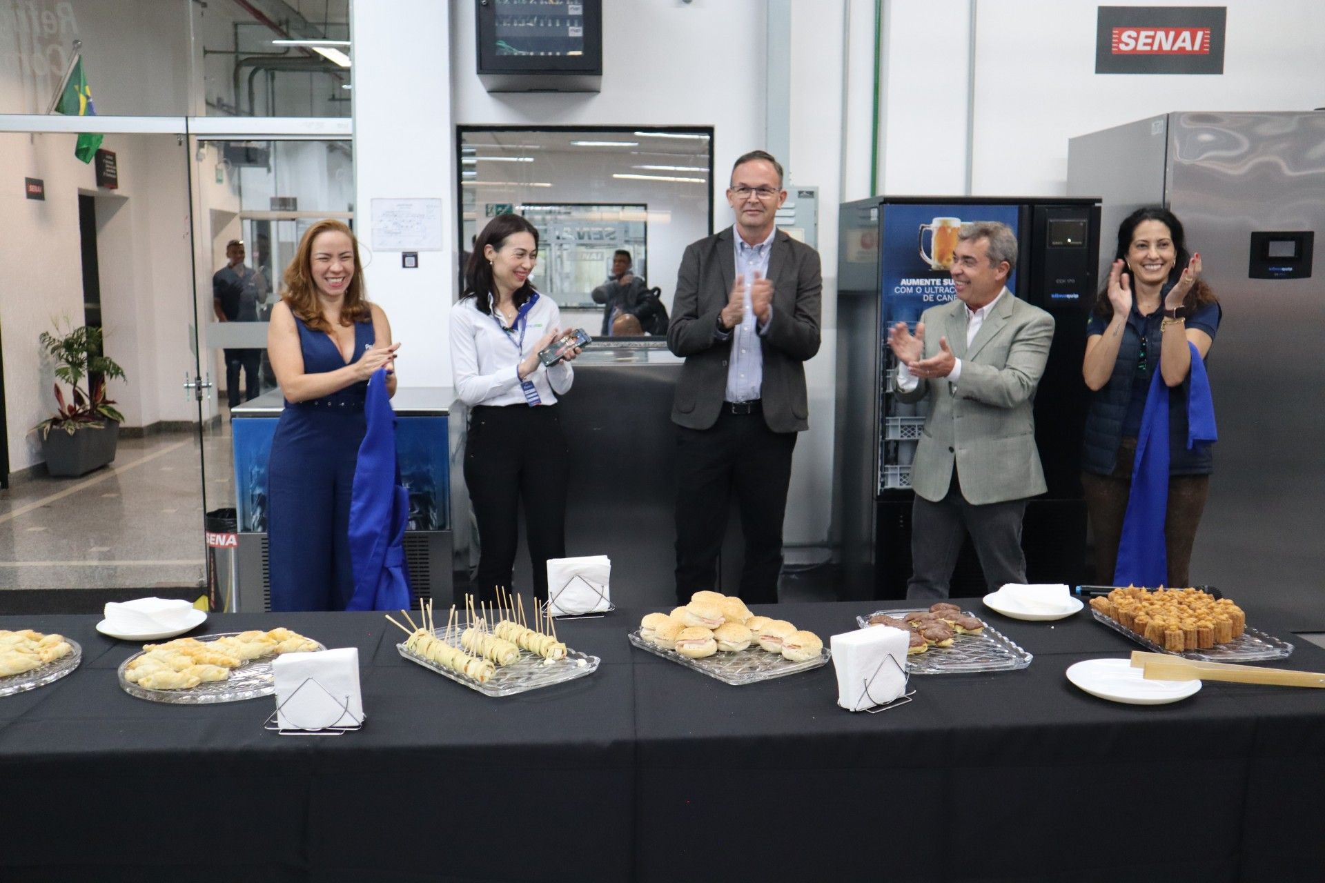 Pessoas aplaudindo em um evento, com lanches sobre uma mesa.