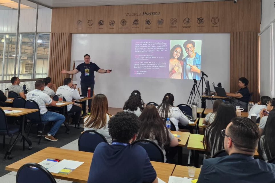 Um homem faz uma apresentação em uma sala de aula. Os alunos sentam-se em carteiras, de frente para o apresentador e para uma tela com uma imagem.