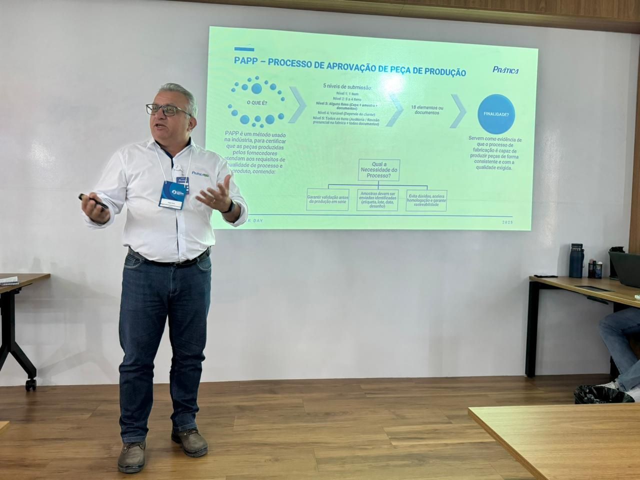 Homem apresentando-se diante de uma tela com um diagrama. Ele gesticula com as mãos e veste camisa branca e calça jeans.