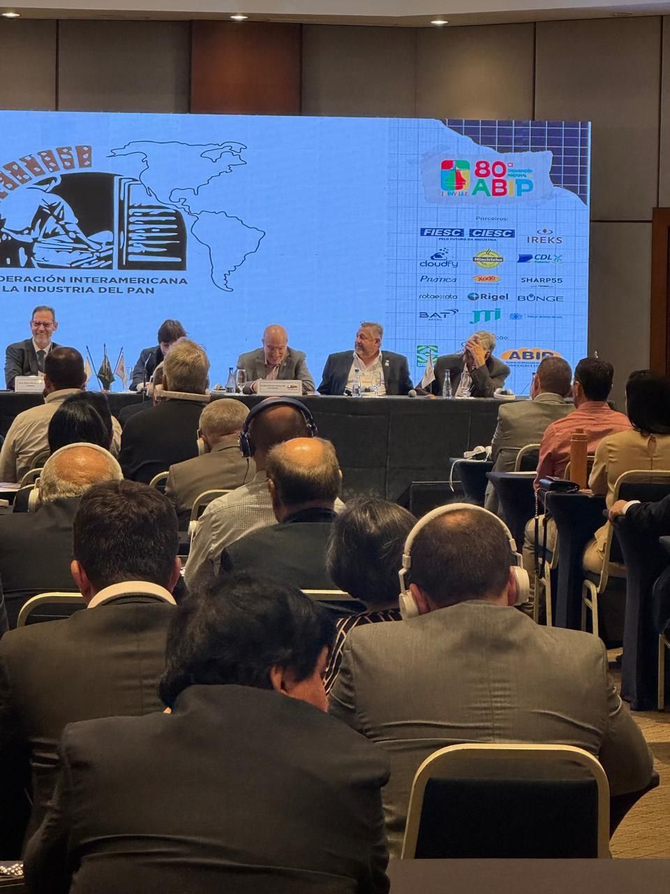 Um grupo de pessoas está sentado em frente a uma tela grande em uma sala de conferências.