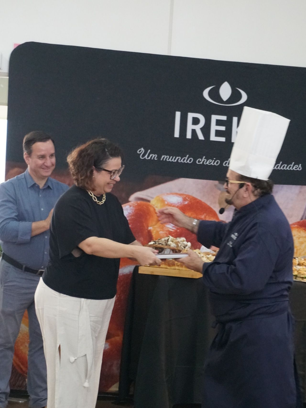 Um chef apresenta comida a uma mulher. Um homem aplaude ao fundo. Logotipo do IREI sobre fundo preto.