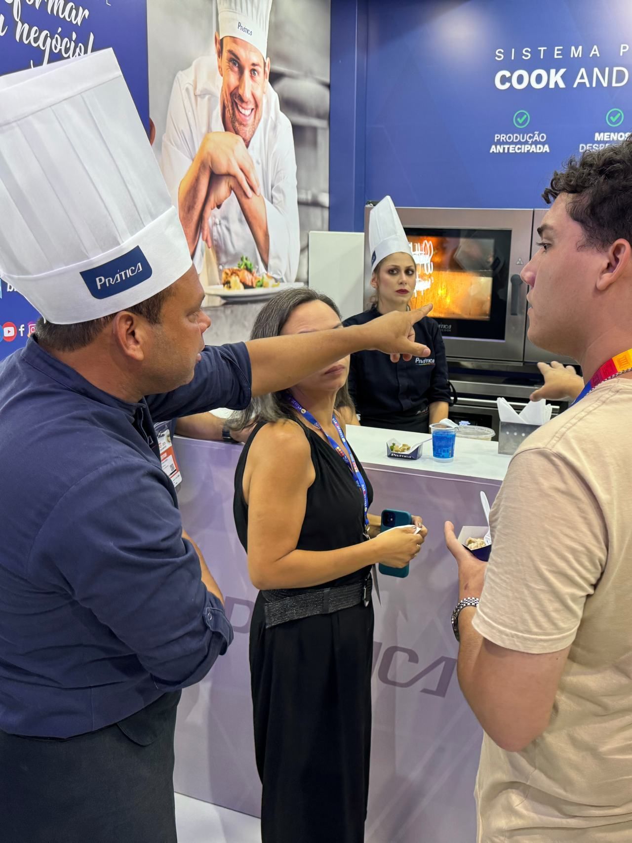 Um homem com chapéu de chef aponta para uma mulher de vestido preto.