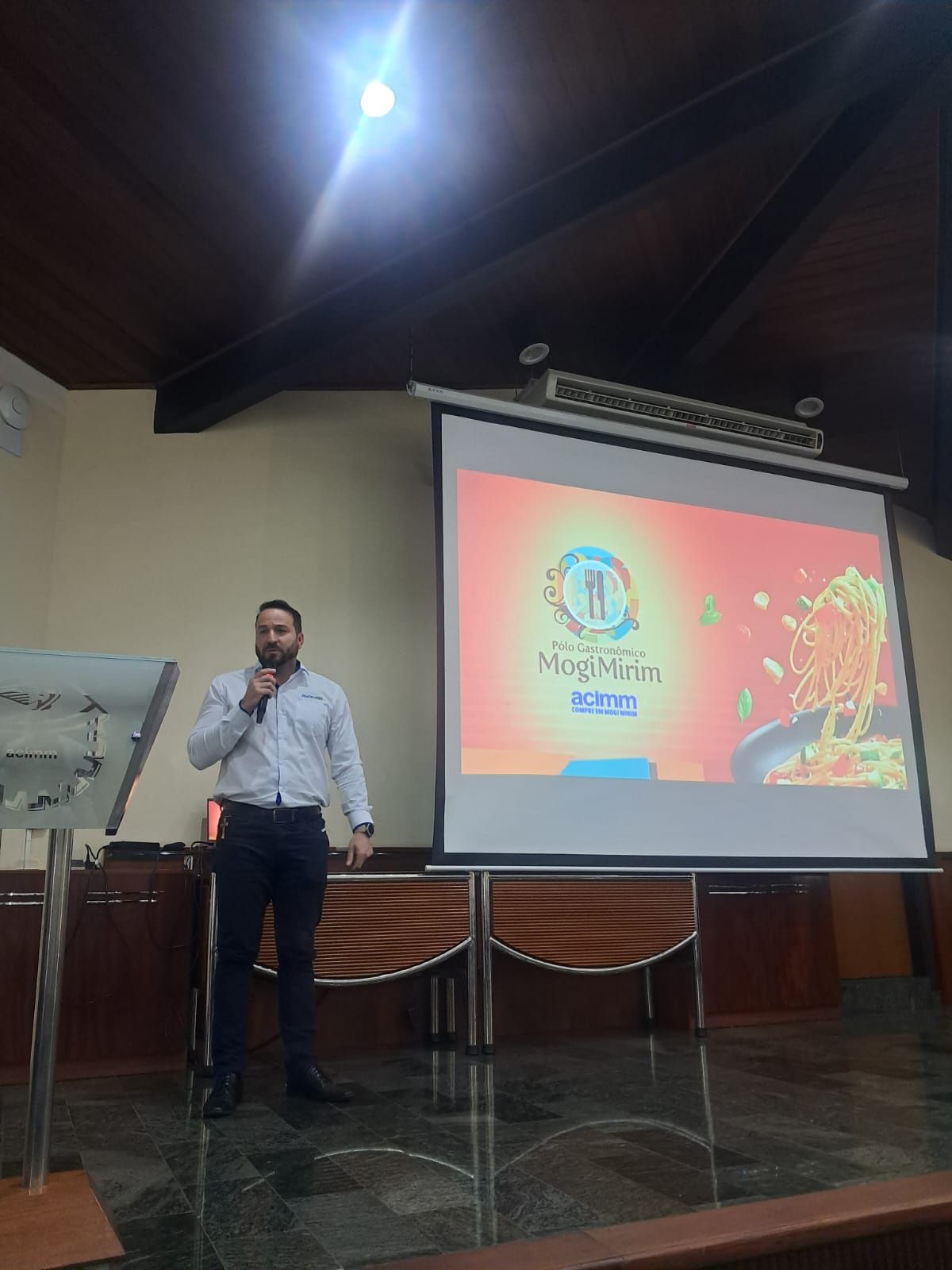 Homem fazendo uma apresentação em frente a uma tela com um logotipo relacionado a alimentos, possivelmente em um auditório.
