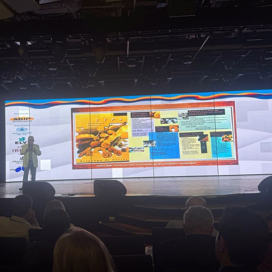 Um homem está fazendo uma apresentação em um palco em frente a uma tela grande.