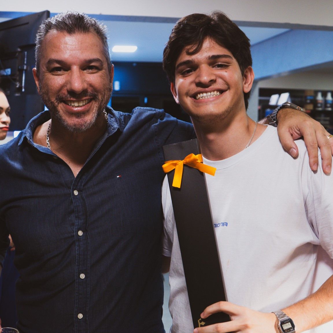 Dois homens estão posando para uma foto e um está segurando uma caixa preta com um laço laranja