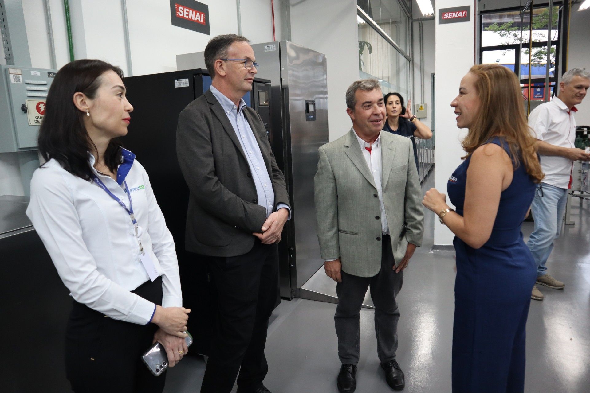 Grupo de pessoas em um laboratório, conversando perto de equipamentos. Mulher de vestido azul gesticulando.