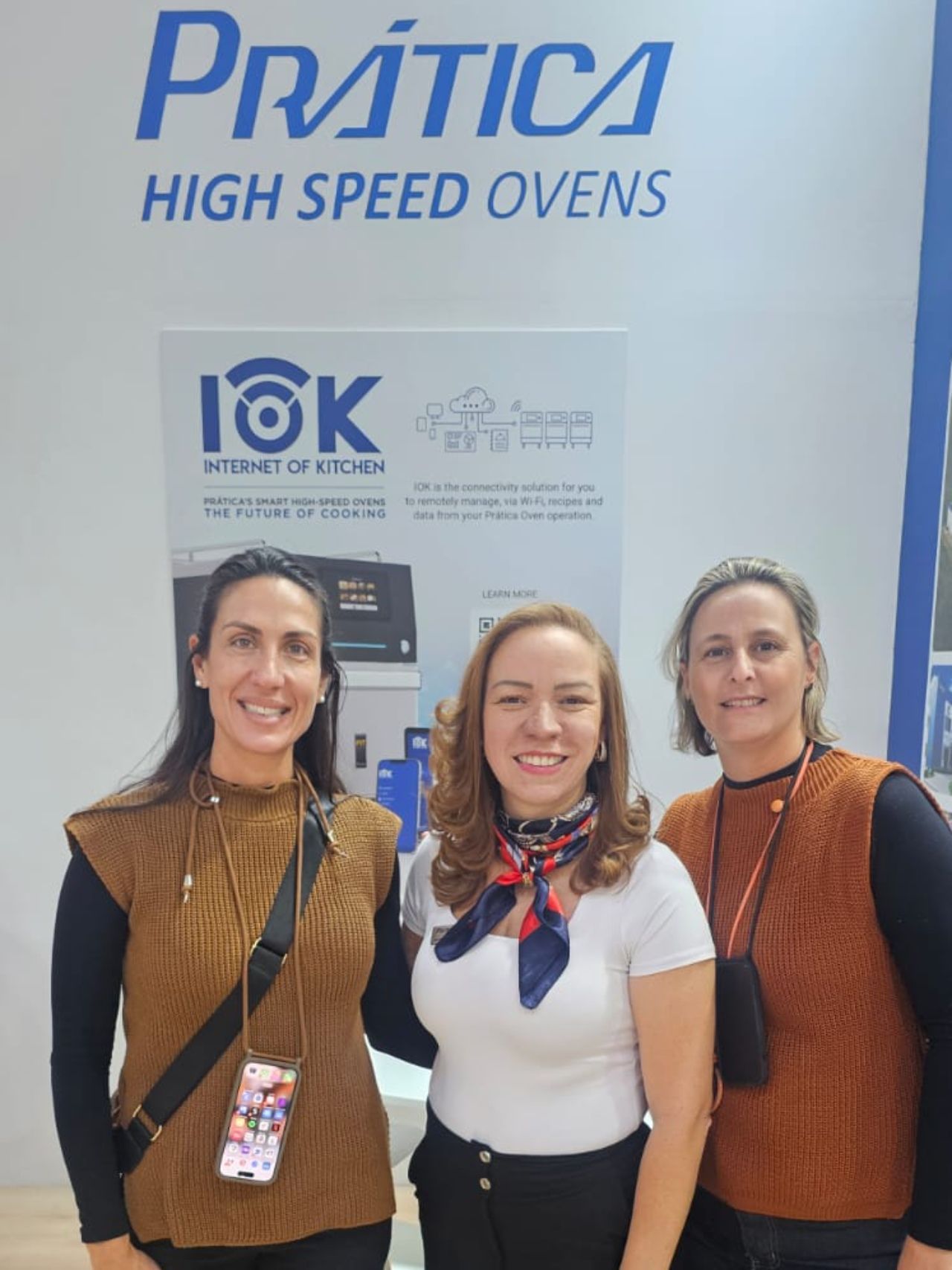 Três mulheres sorrindo em frente a uma exposição de fornos de alta velocidade Pratica.