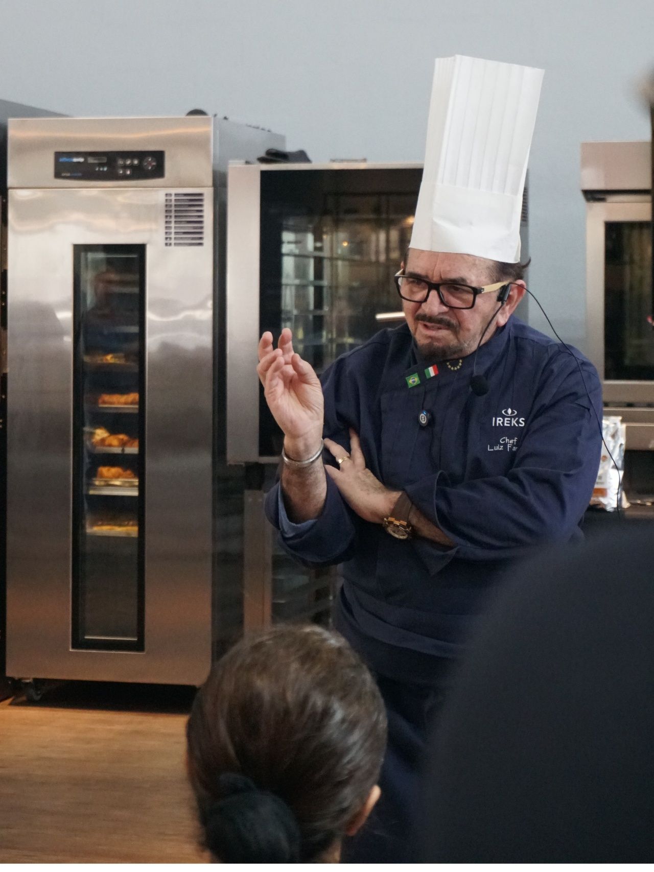 Chef de chapéu alto gesticulando em frente aos fornos, dando uma aula.