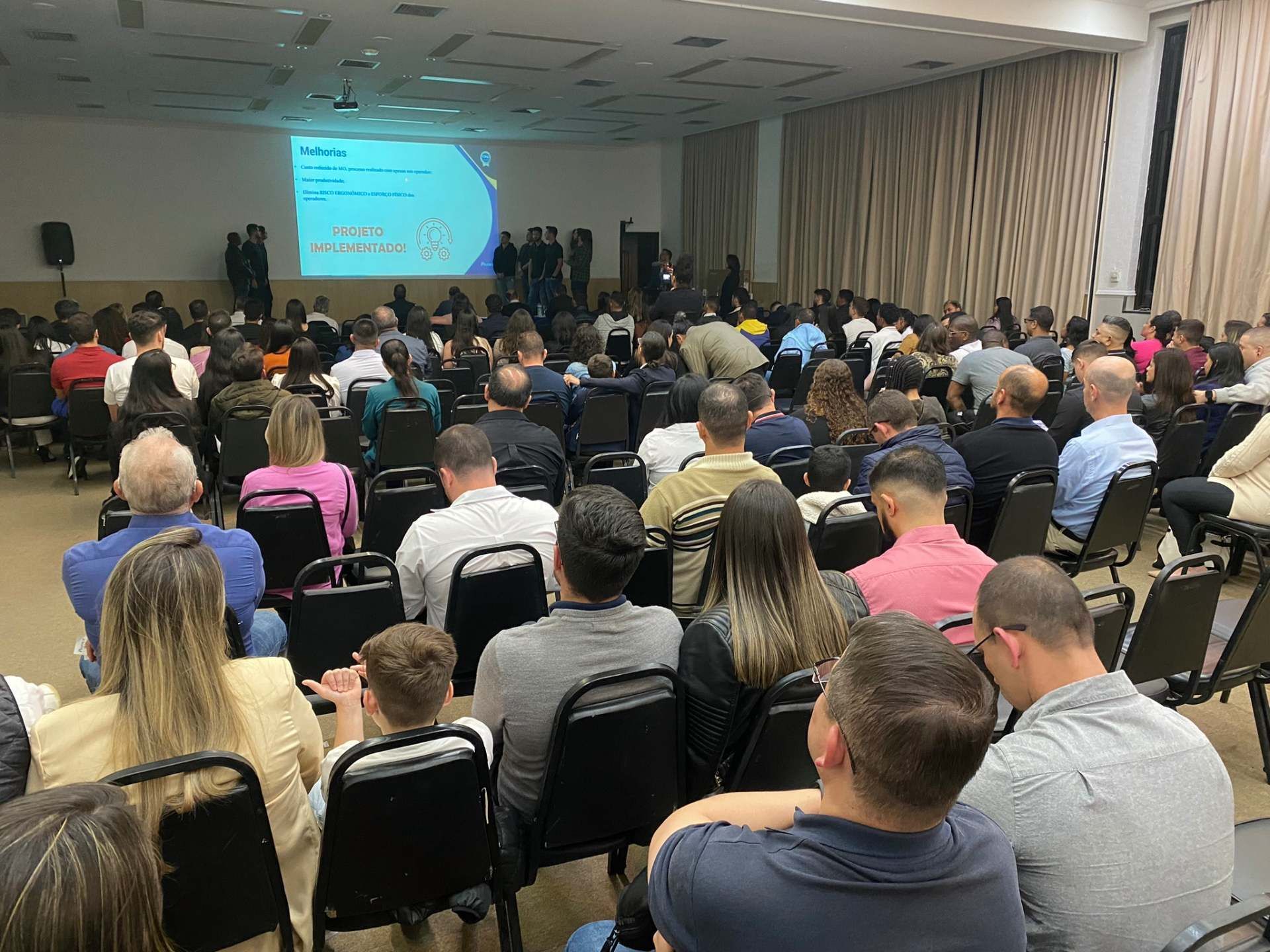 Um grande grupo de pessoas está sentado em um grande auditório assistindo a uma apresentação.