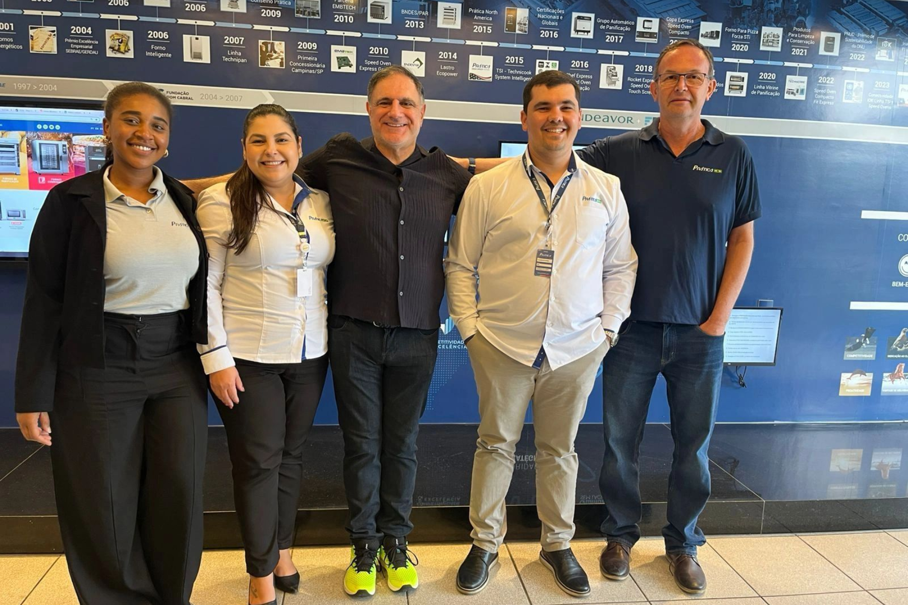 Cinco pessoas sorrindo, posando juntas dentro de um prédio. Atrás delas, um muro azul-marinho.