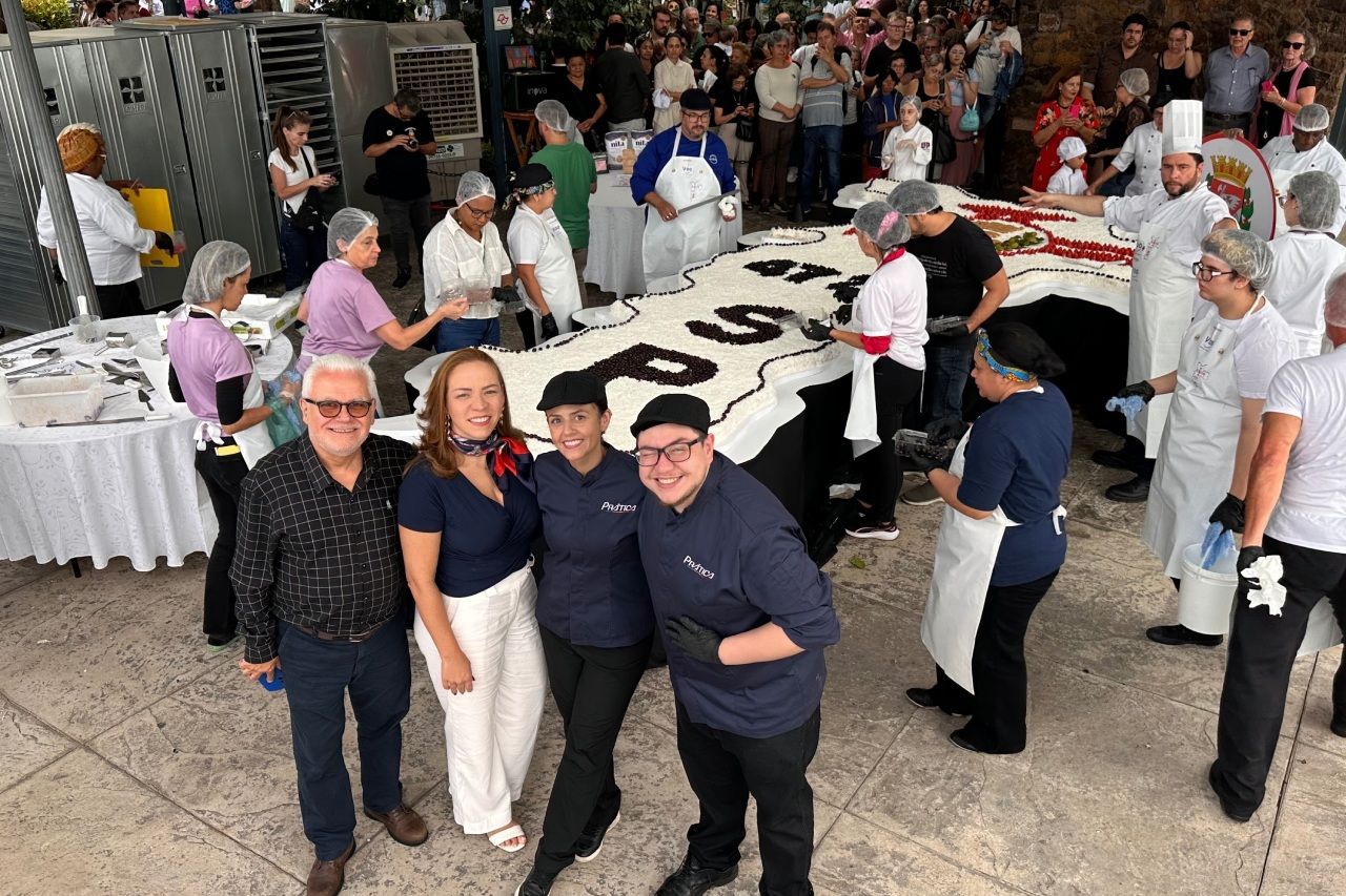 Pessoas ao redor de um grande bolo decorado; algumas usando chapéus e aventais de chef, outras posando para uma foto. Ao ar livre.