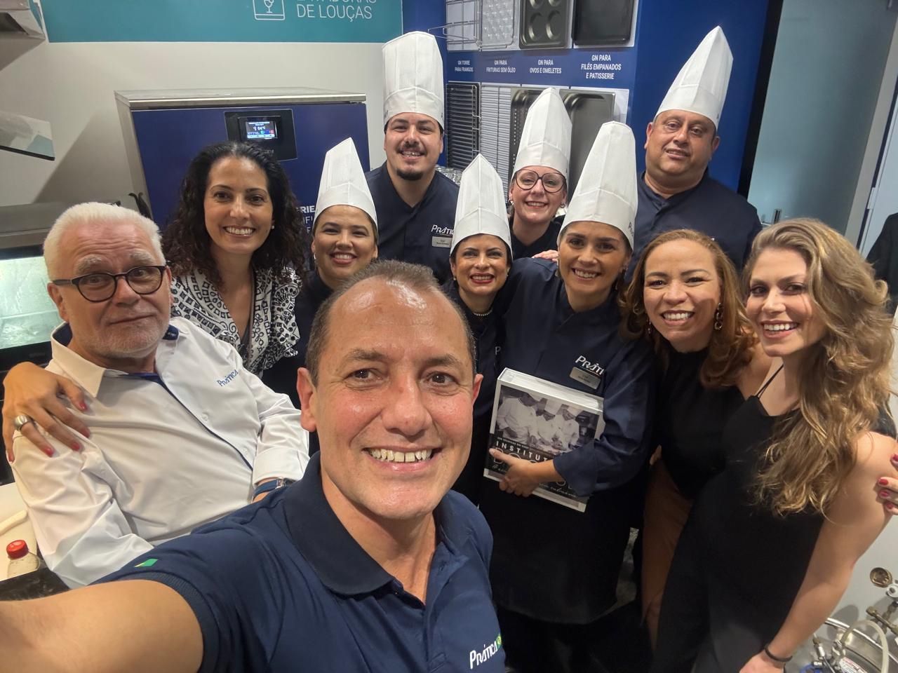 Grupo de pessoas, algumas com chapéus de chef, sorrindo e tirando uma selfie. Fundo azul e branco.