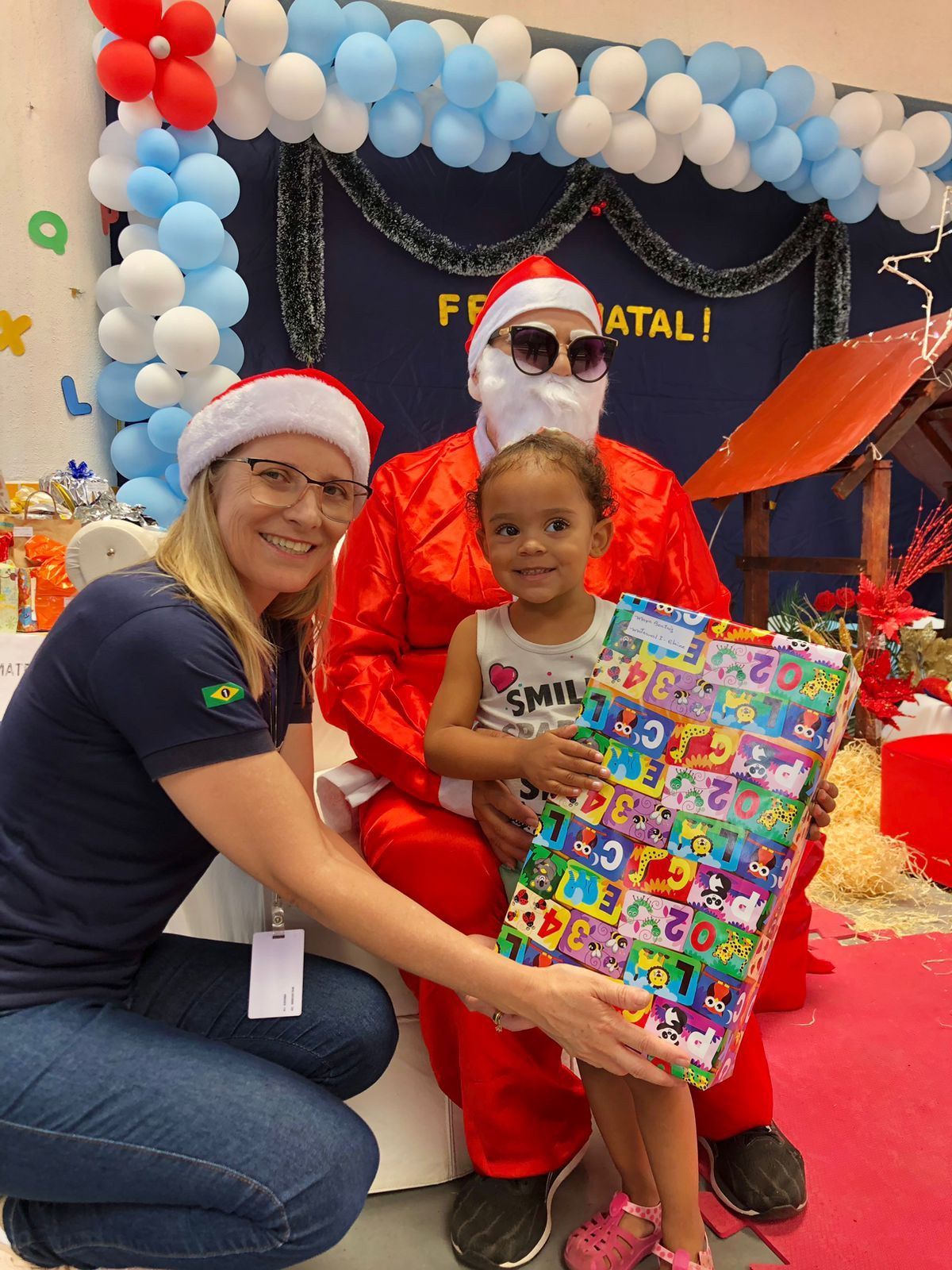 Uma mulher está ajoelhada ao lado de uma menina segurando uma caixa de presente enquanto está sentada no colo do Papai Noel.