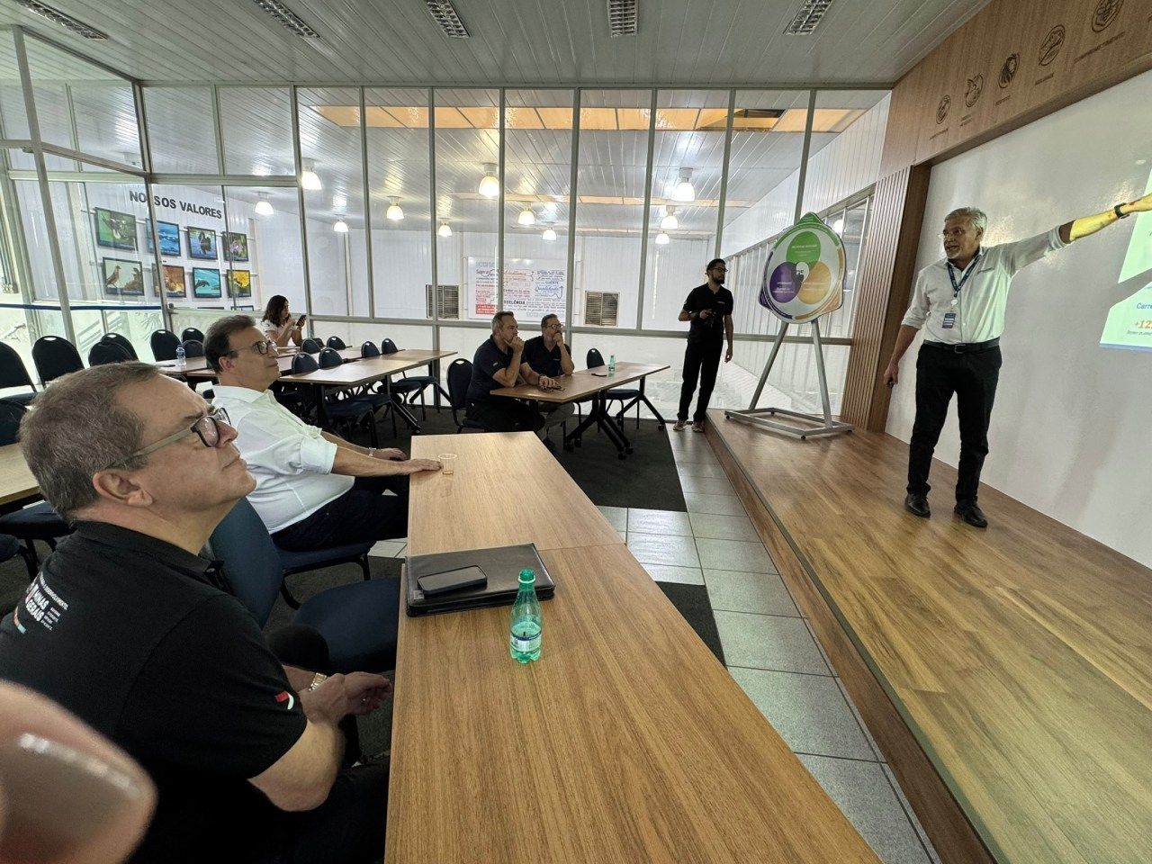 Um grupo de pessoas está sentado em mesas em uma sala enquanto um homem está em um palco fazendo uma apresentação.