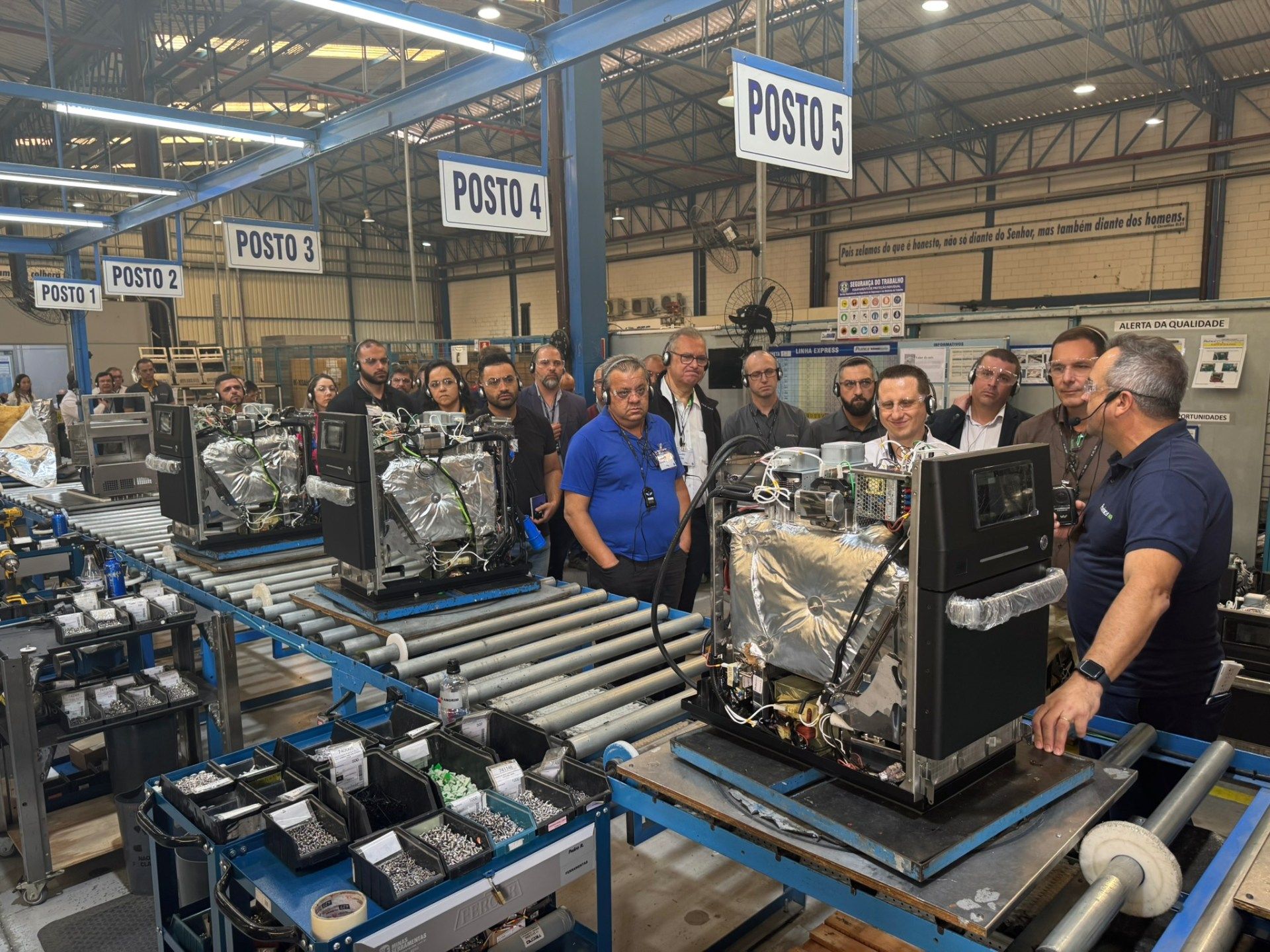 Um grupo de pessoas está em pé ao redor de uma correia transportadora em uma fábrica.