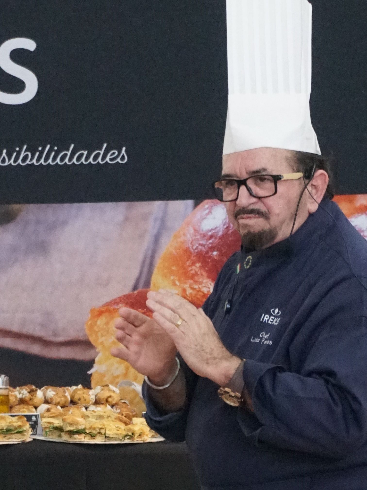 Chef batendo palmas com chapéu alto, óculos e jaqueta azul escura, perto de uma exposição de doces.