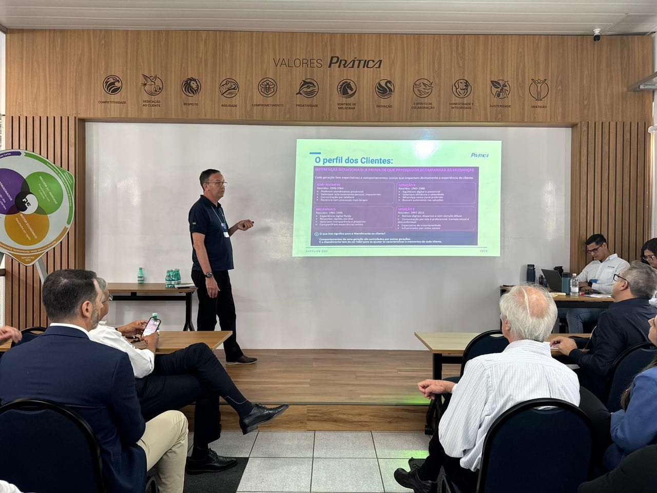 Um homem apresenta uma apresentação de slides para uma plateia sentada. A apresentação acontece em uma sala branca.