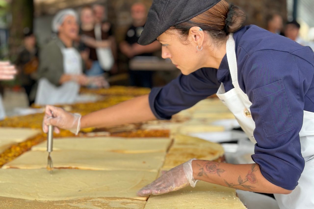 Mulher de touca e avental pincelando um bolo em uma mesa comprida ao ar livre.