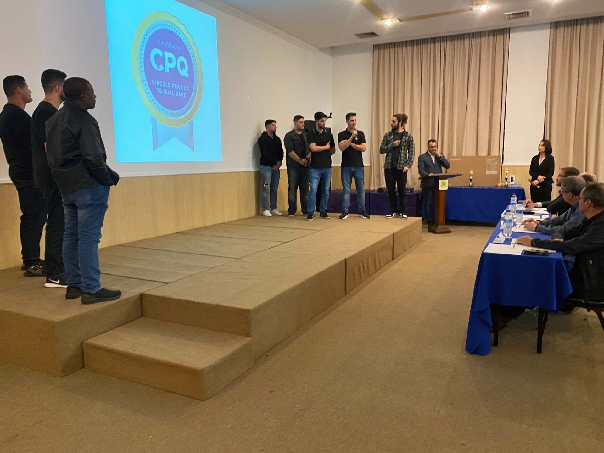 Um grupo de pessoas em pé em um palco em frente a uma tela de projetor que diz cpq