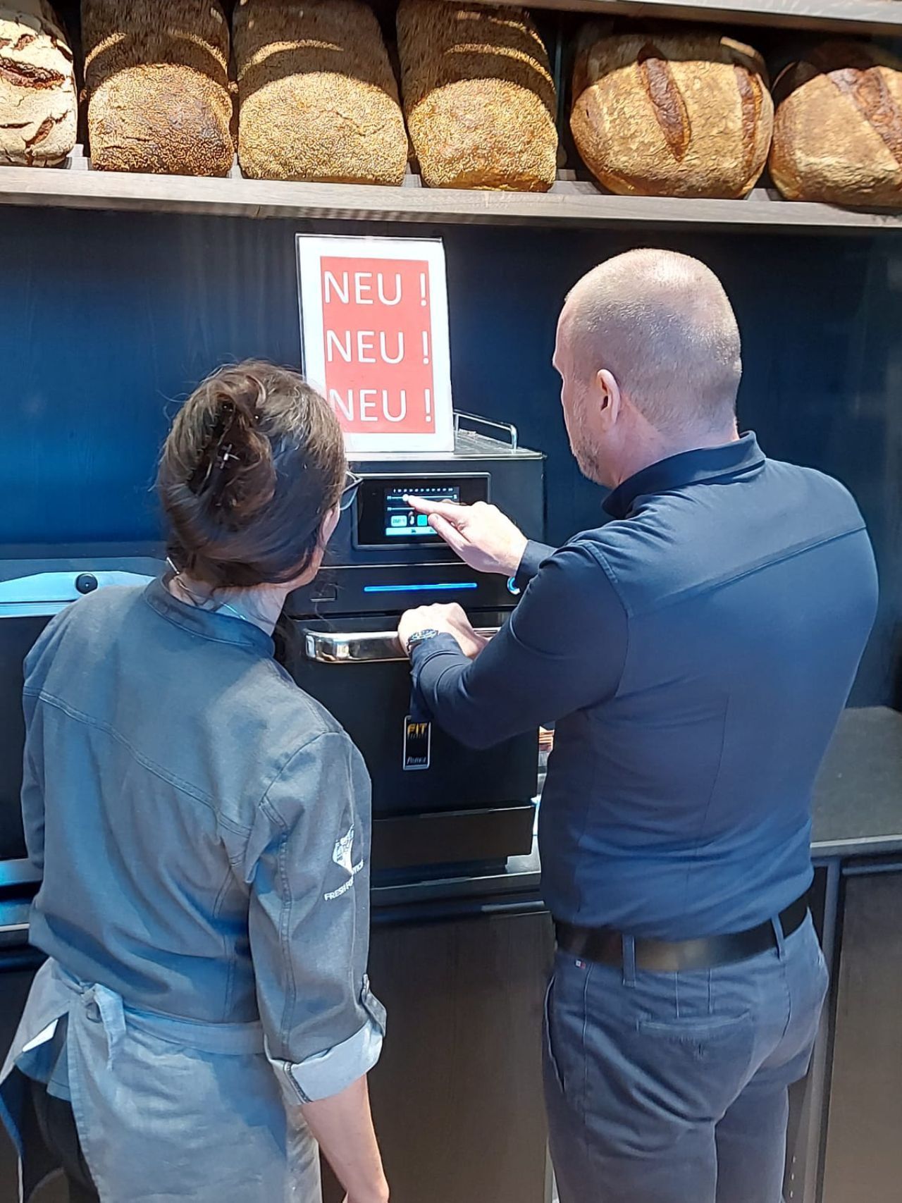 Um homem e uma mulher estão olhando para uma máquina em uma padaria.