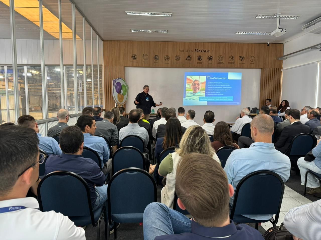 Um homem faz uma apresentação para uma plateia sentada em uma sala com uma tela.