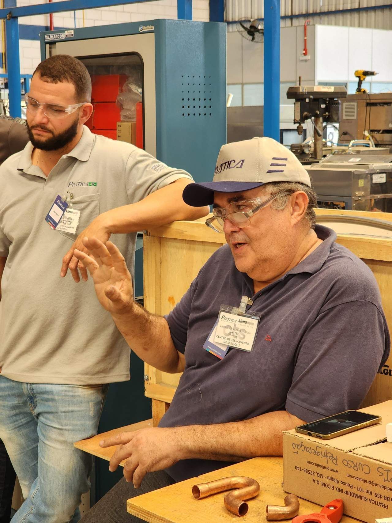Dois homens em uma fábrica, um usando um chapéu que diz