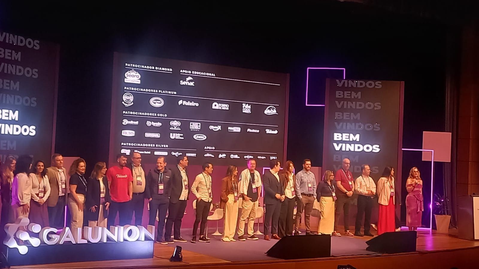 Grupo de pessoas no palco de um evento, com uma tela exibindo logotipos e o texto