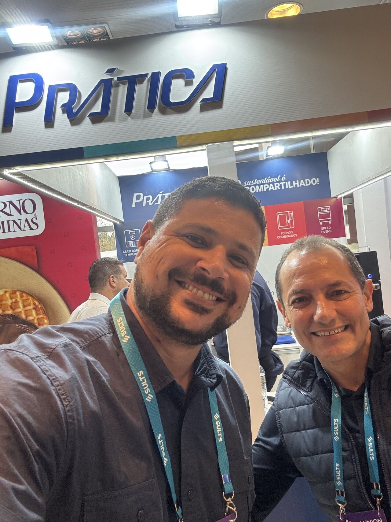 Dois homens sorriem, tirando uma selfie em frente ao estande da