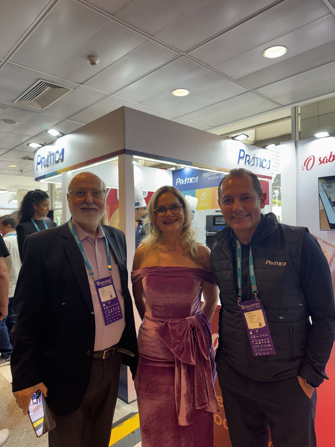 Três pessoas posam em frente a um estande. Uma mulher de vestido rosa está entre dois homens. O estande ostenta o logotipo