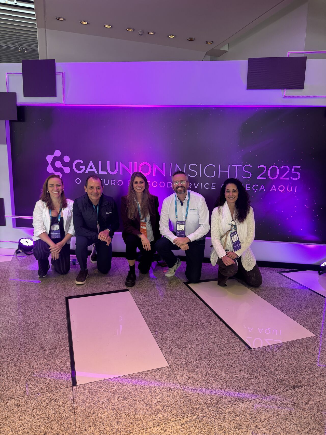 Cinco pessoas ajoelhadas no palco de uma conferência. Fundo roxo com logotipo.