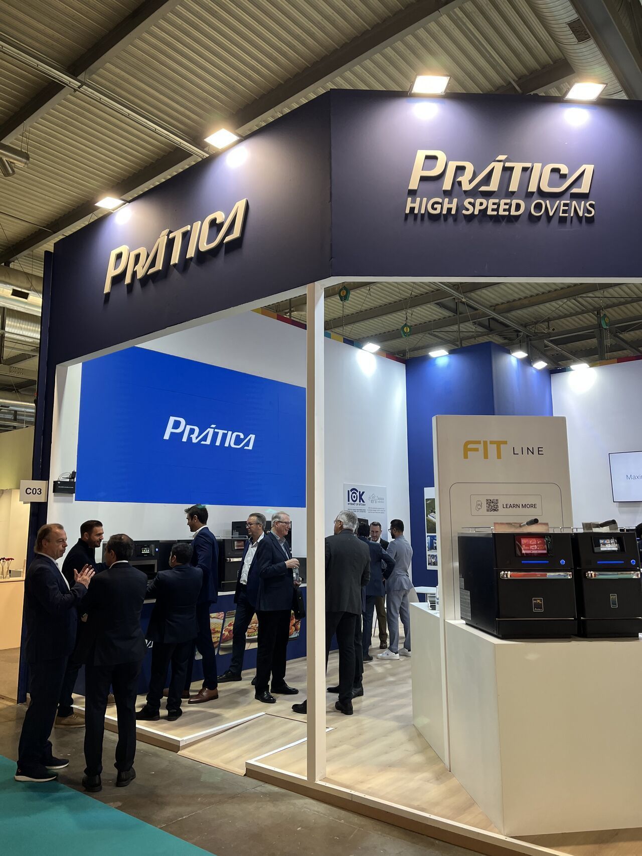 Estande da Prática High Speed ​​Ovens em uma feira. Pessoas de terno estão no estande, que tem a marca azul e branca.