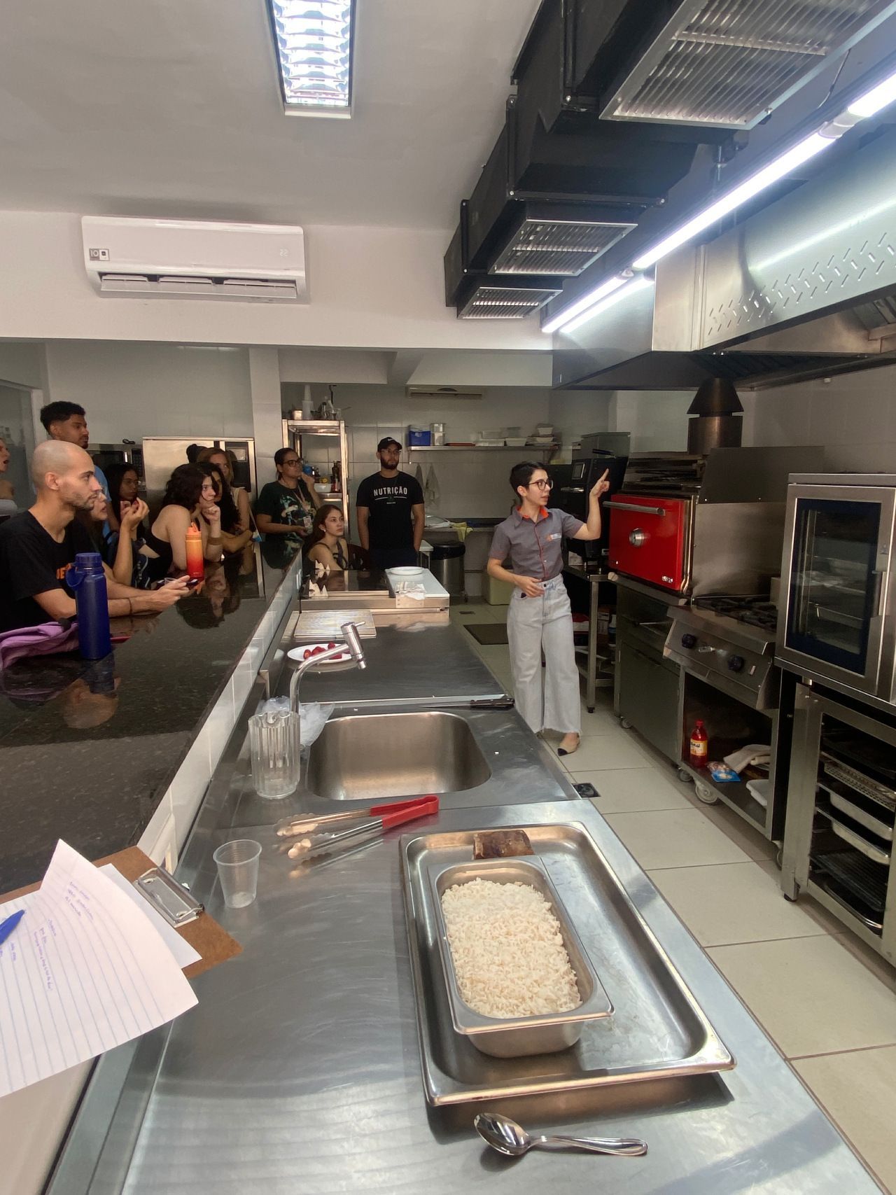 Um grupo de pessoas está de pé ao redor de um longo balcão em uma cozinha.