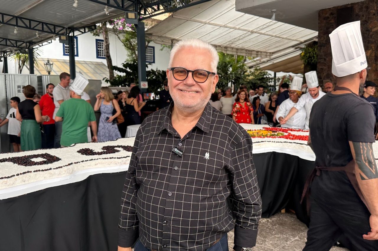 Homem de óculos sorri em frente a um bolo gigante, rodeado de pessoas e chefs. Evento ao ar livre.