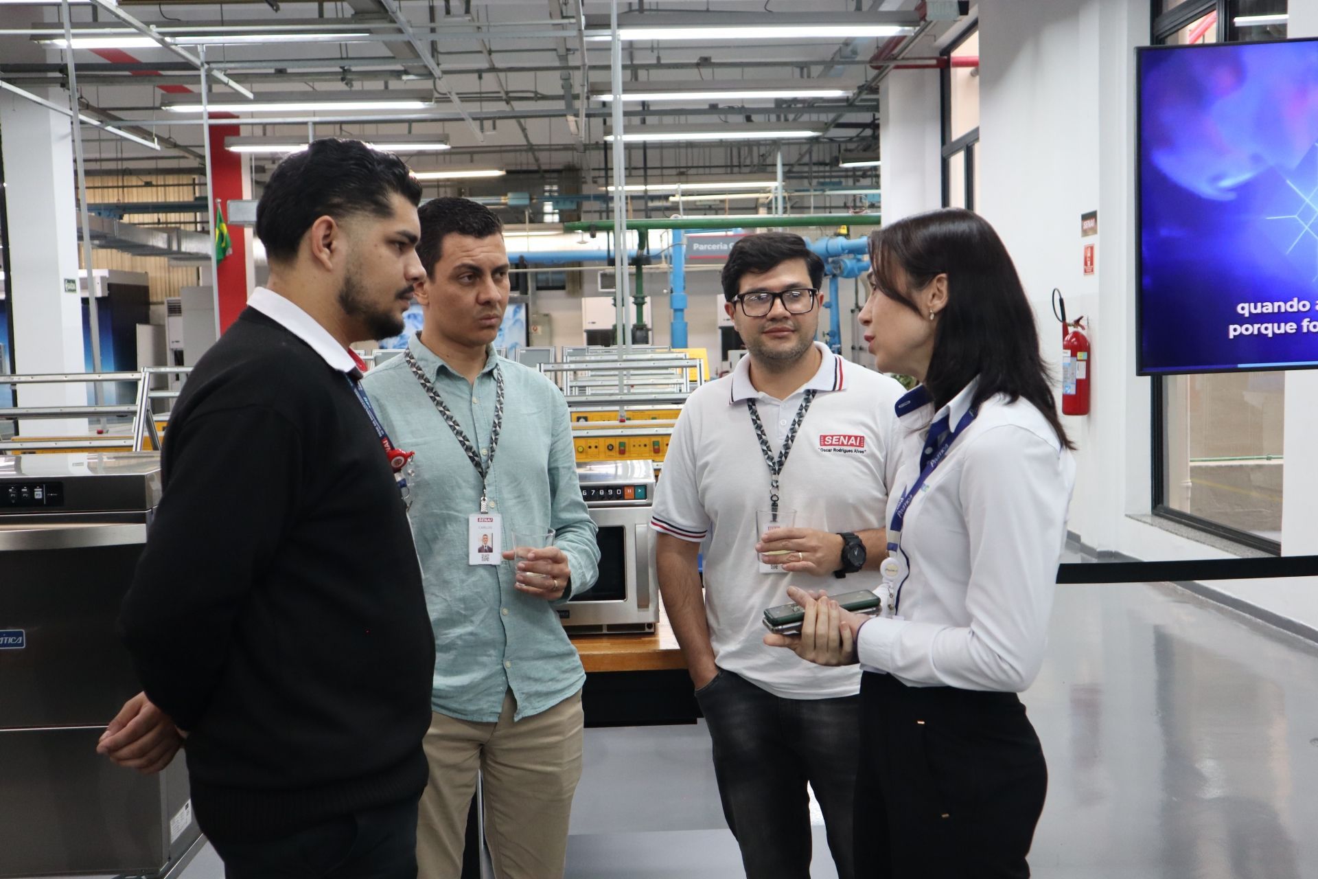 Quatro pessoas conversando em um ambiente fabril. Uma mulher uniformizada gesticula enquanto fala com três homens.
