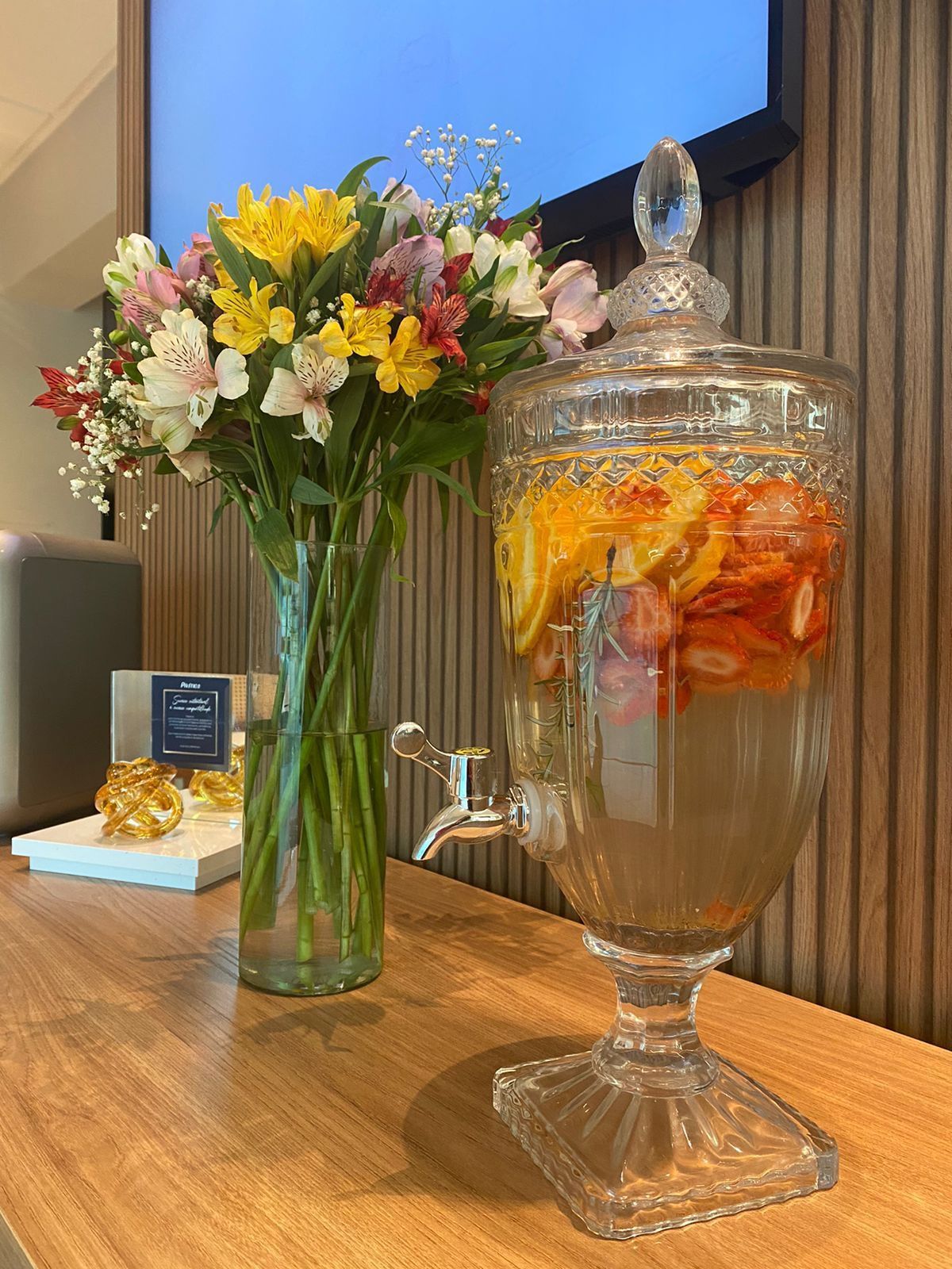 Um dispensador de vidro cheio de frutas e flores está sobre uma mesa de madeira ao lado de um vaso de flores.