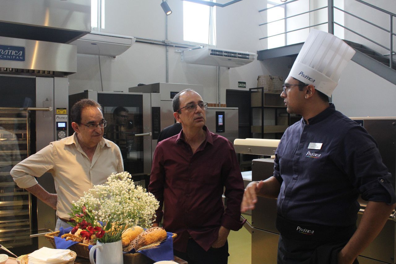Um homem usando um chapéu de chef está conversando com outros dois homens em uma cozinha