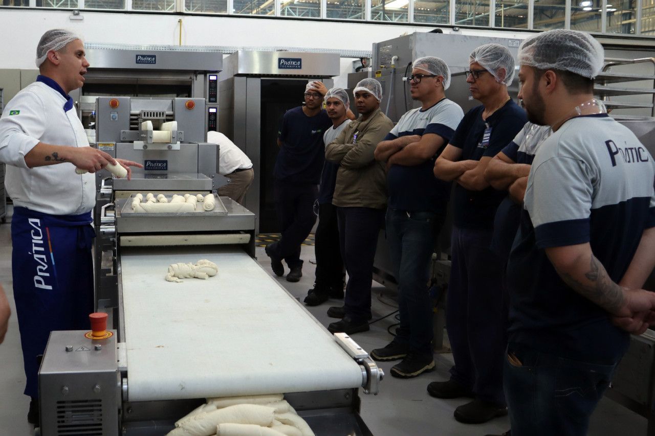 Um grupo de homens está em pé ao redor de uma esteira rolante em uma padaria.