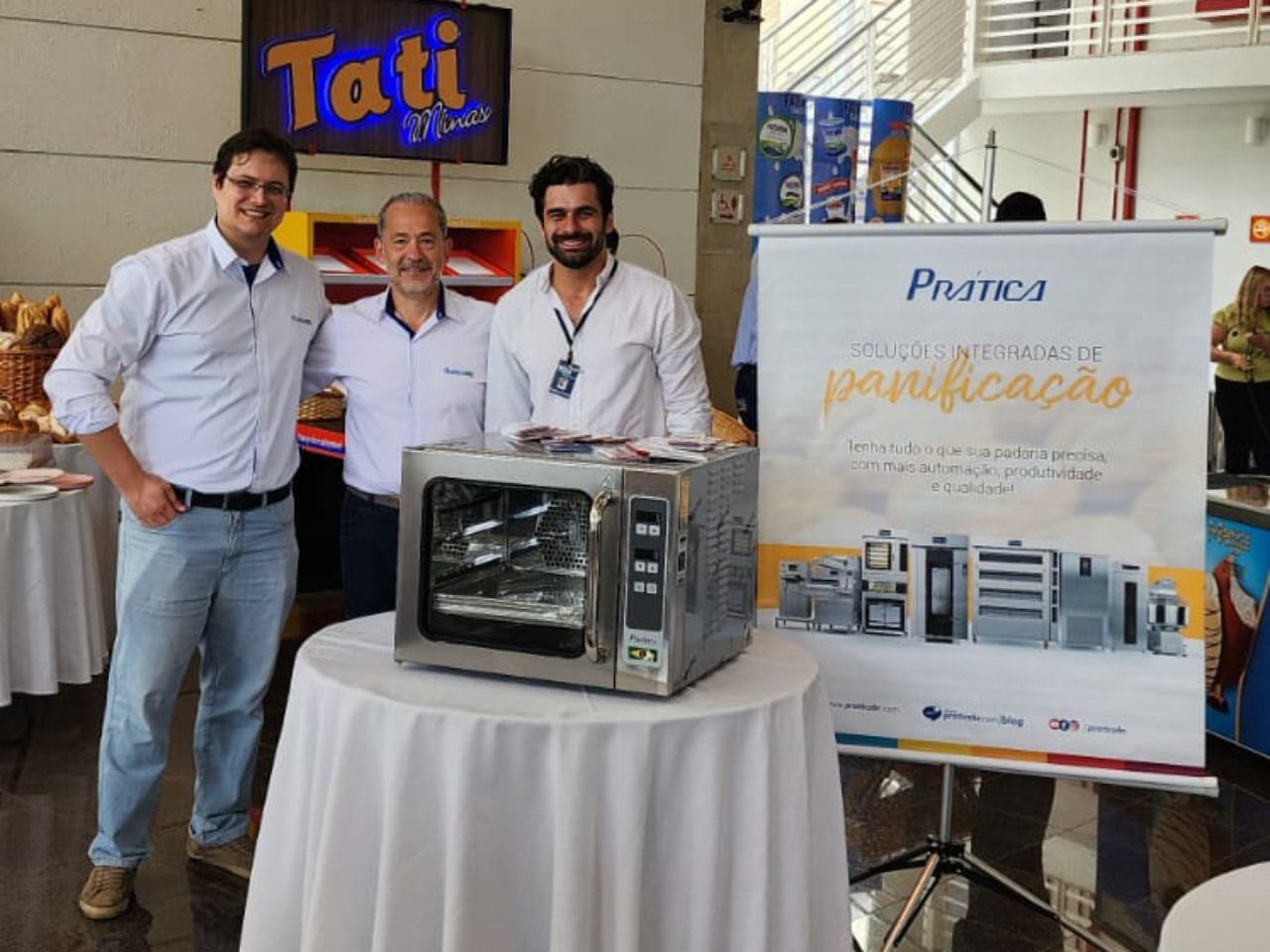 Três homens posam para uma foto em frente a uma placa que diz tati