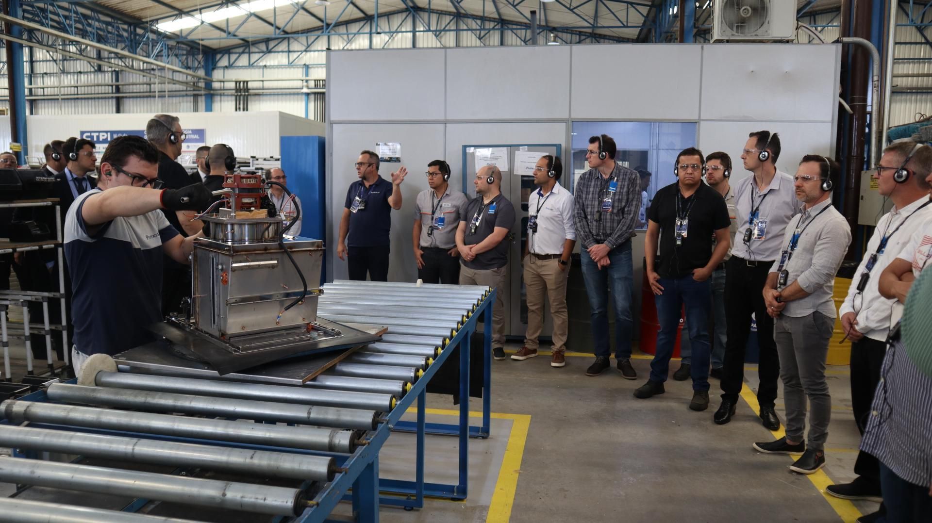 Um grupo de homens está em pé ao redor de uma correia transportadora em uma fábrica.