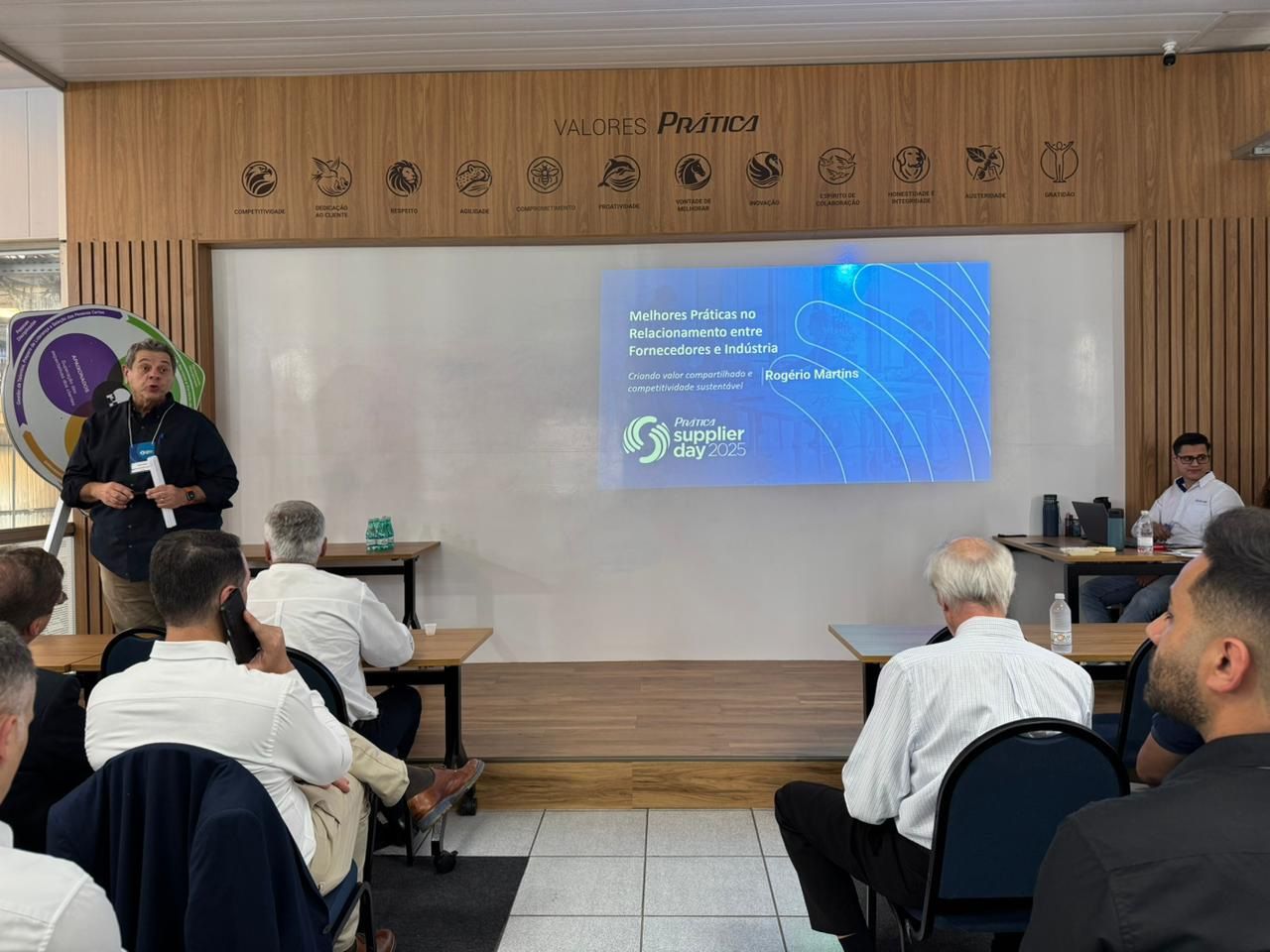 Um apresentador faz uma palestra em uma conferência, com a plateia sentada, tela com título, sala com detalhes em madeira.