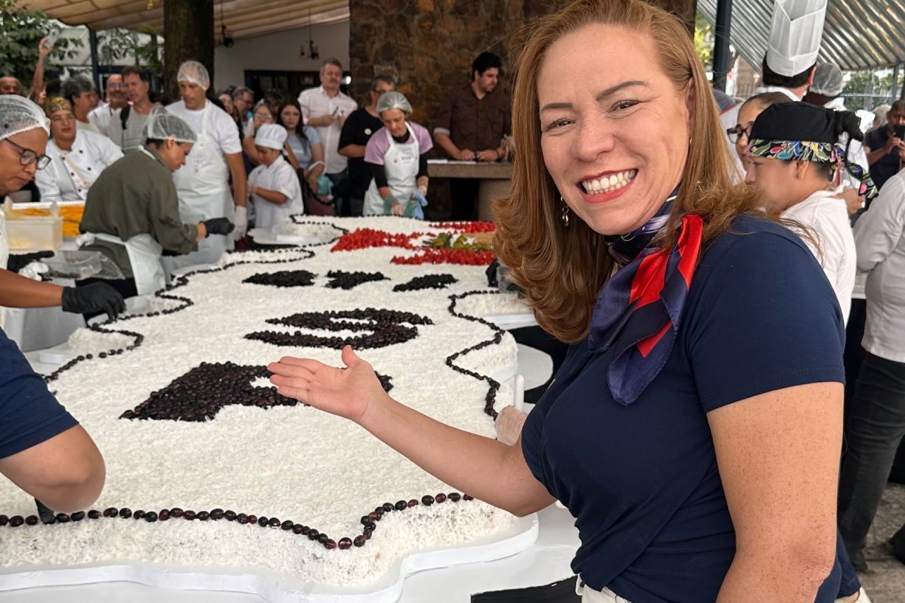 Uma mulher sorri e aponta para um grande bolo decorado com ingredientes em preto e branco. Pessoas trabalham no bolo, com uma multidão ao fundo.
