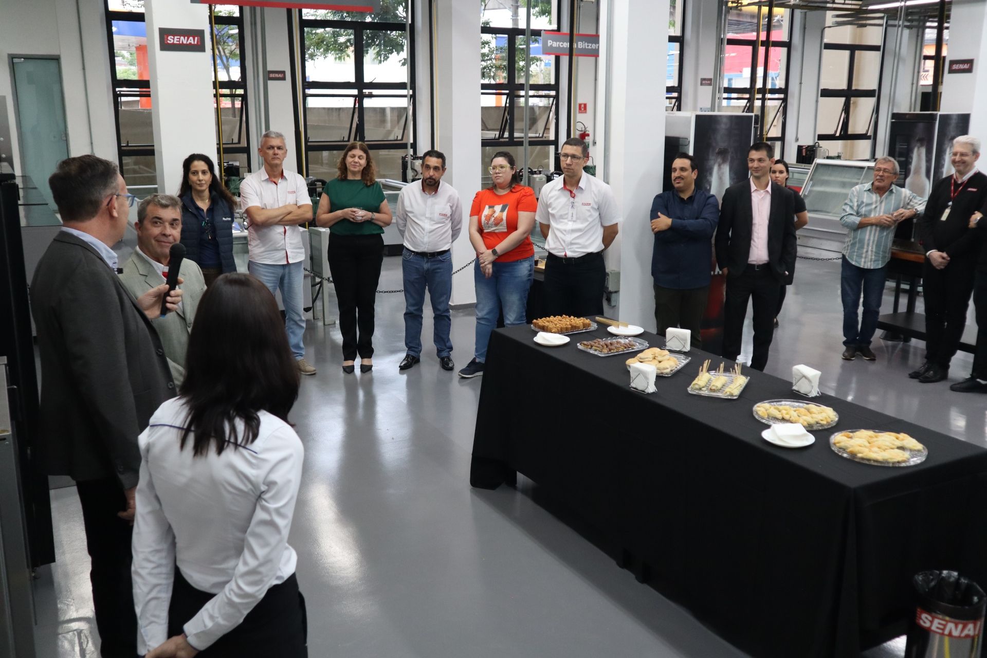 Grupo de pessoas em uma área aberta, algumas olhando em direção à câmera, com uma mesa com lanches em primeiro plano.