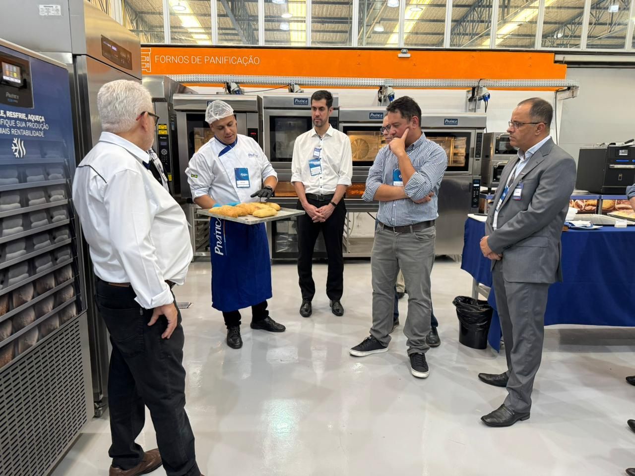 Um grupo de homens em uma exposição de padaria; um chef segura uma bandeja de pão, um homem gesticula.
