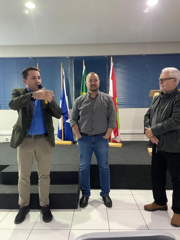 Três homens estão de pé, um ao lado do outro, em uma sala com bandeiras ao fundo.