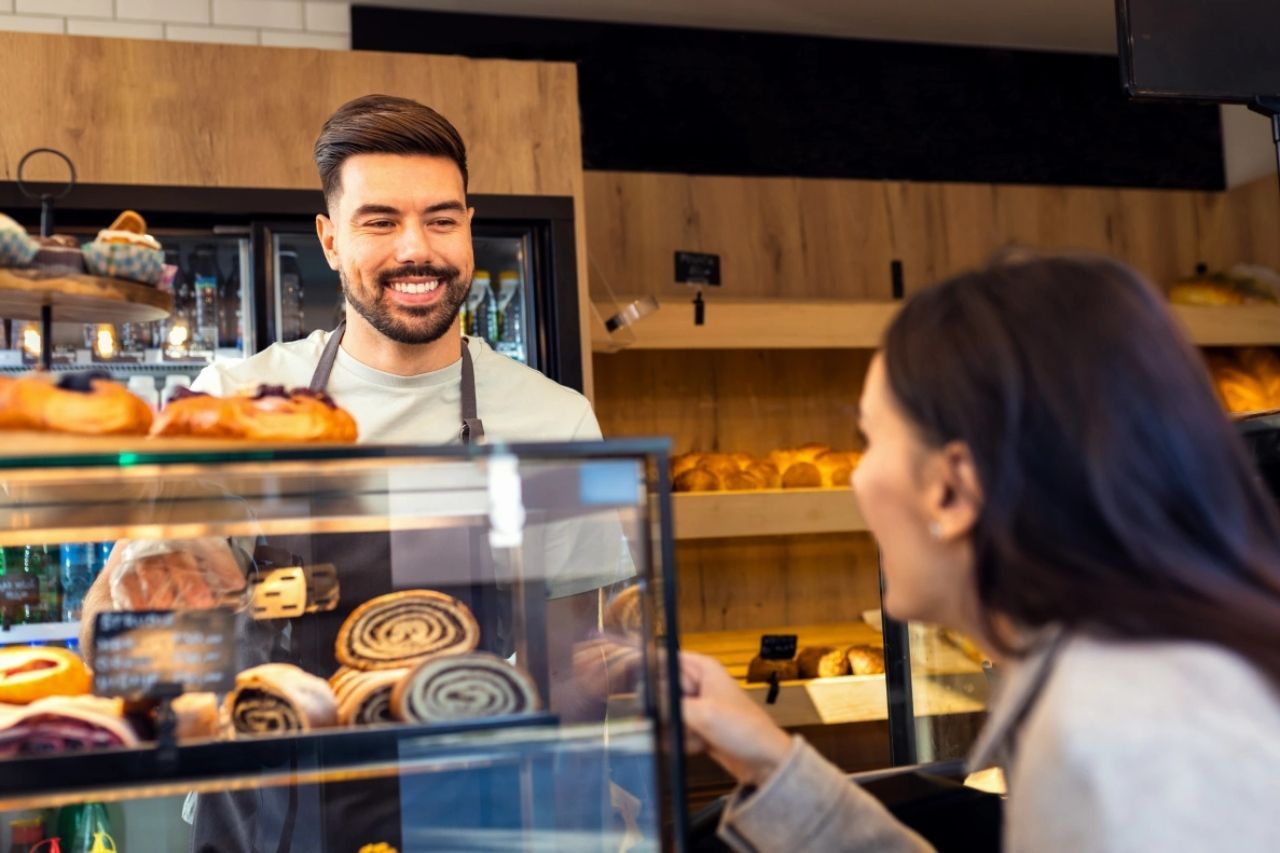 Funcionário sorridente da padaria atendendo uma cliente e uma vitrine de vidro repleta de doces.