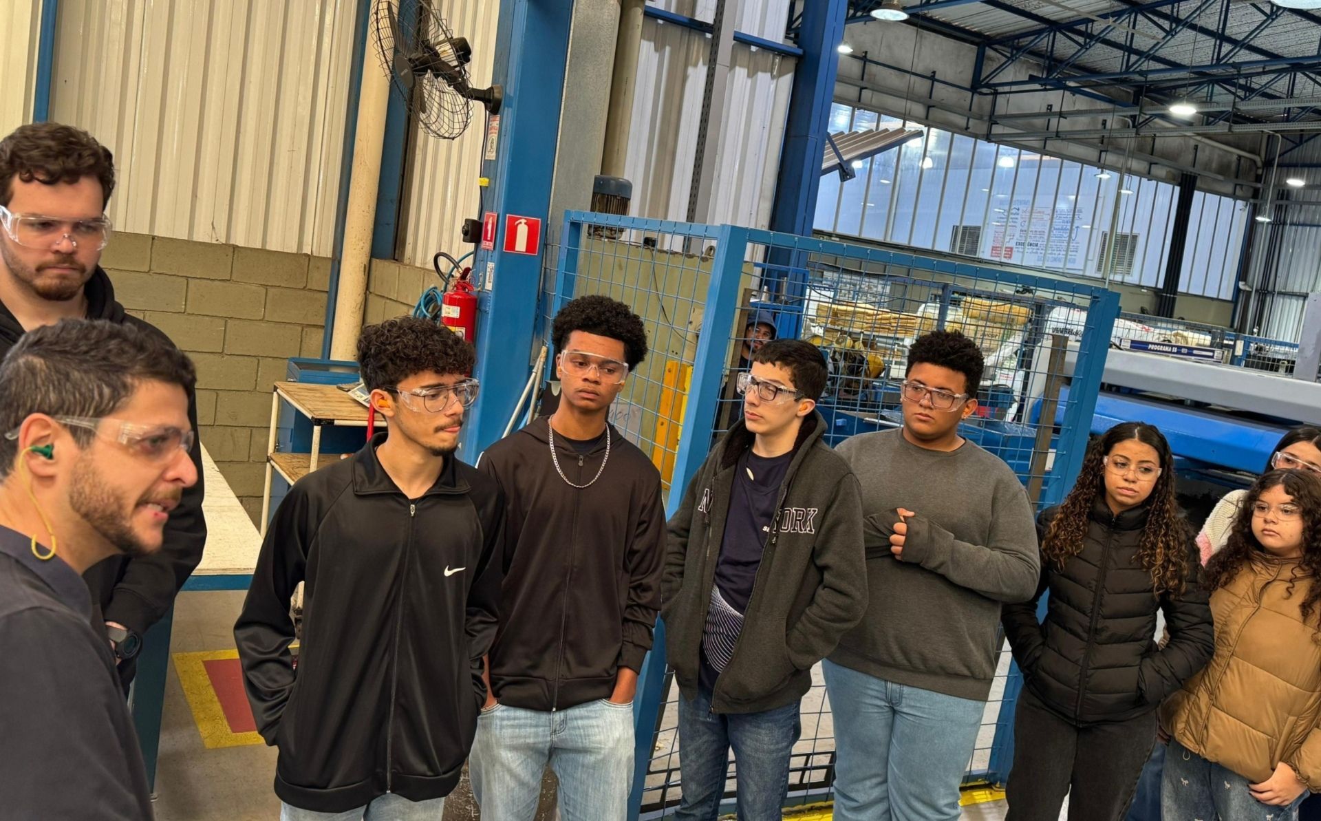 Grupo de estudantes ouve um homem em uma oficina; equipamentos de fábrica ao fundo.