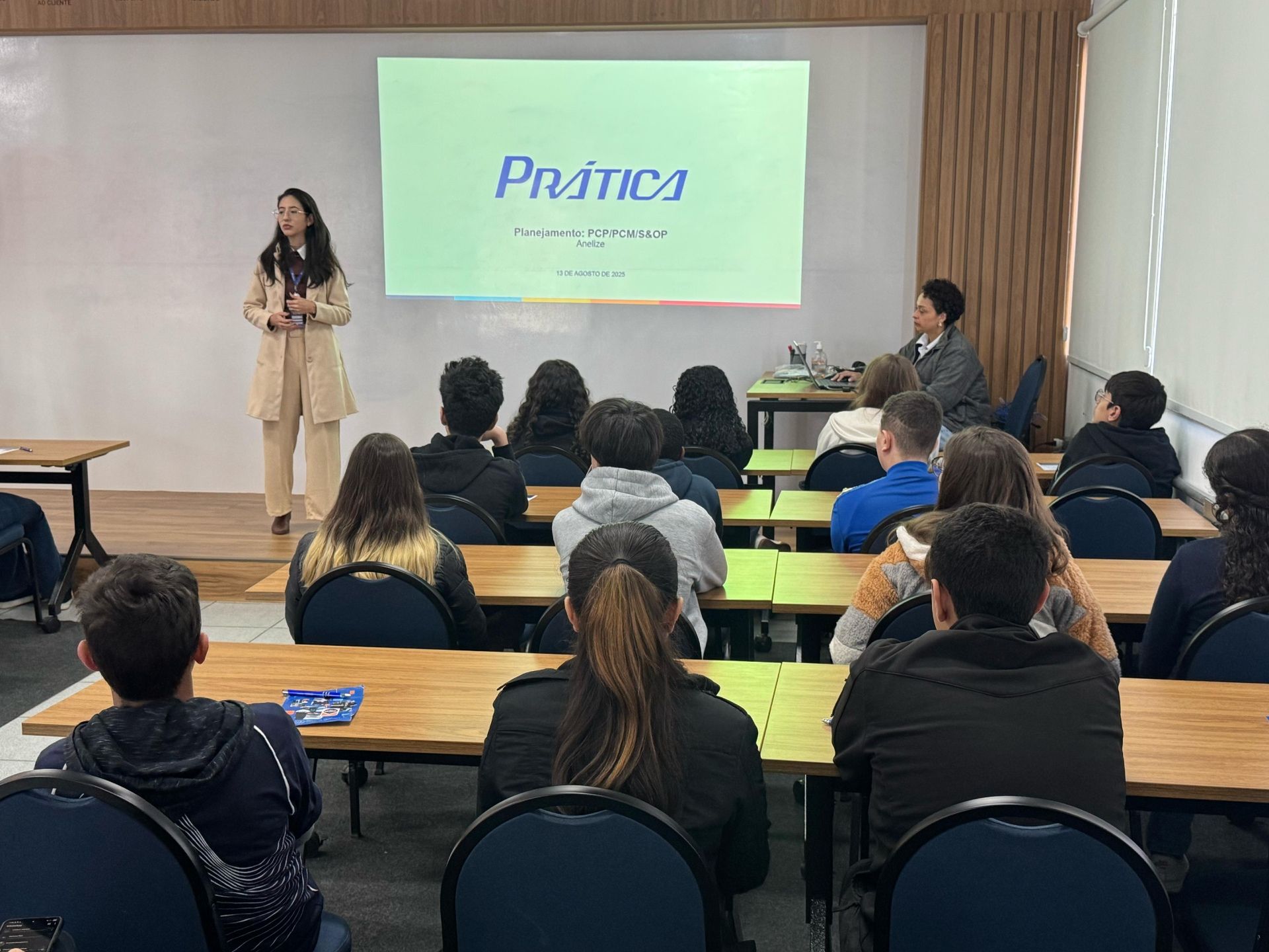 Mulher fazendo apresentação em sala de aula, alunos sentados em carteiras, tela de projeção com a palavra 
