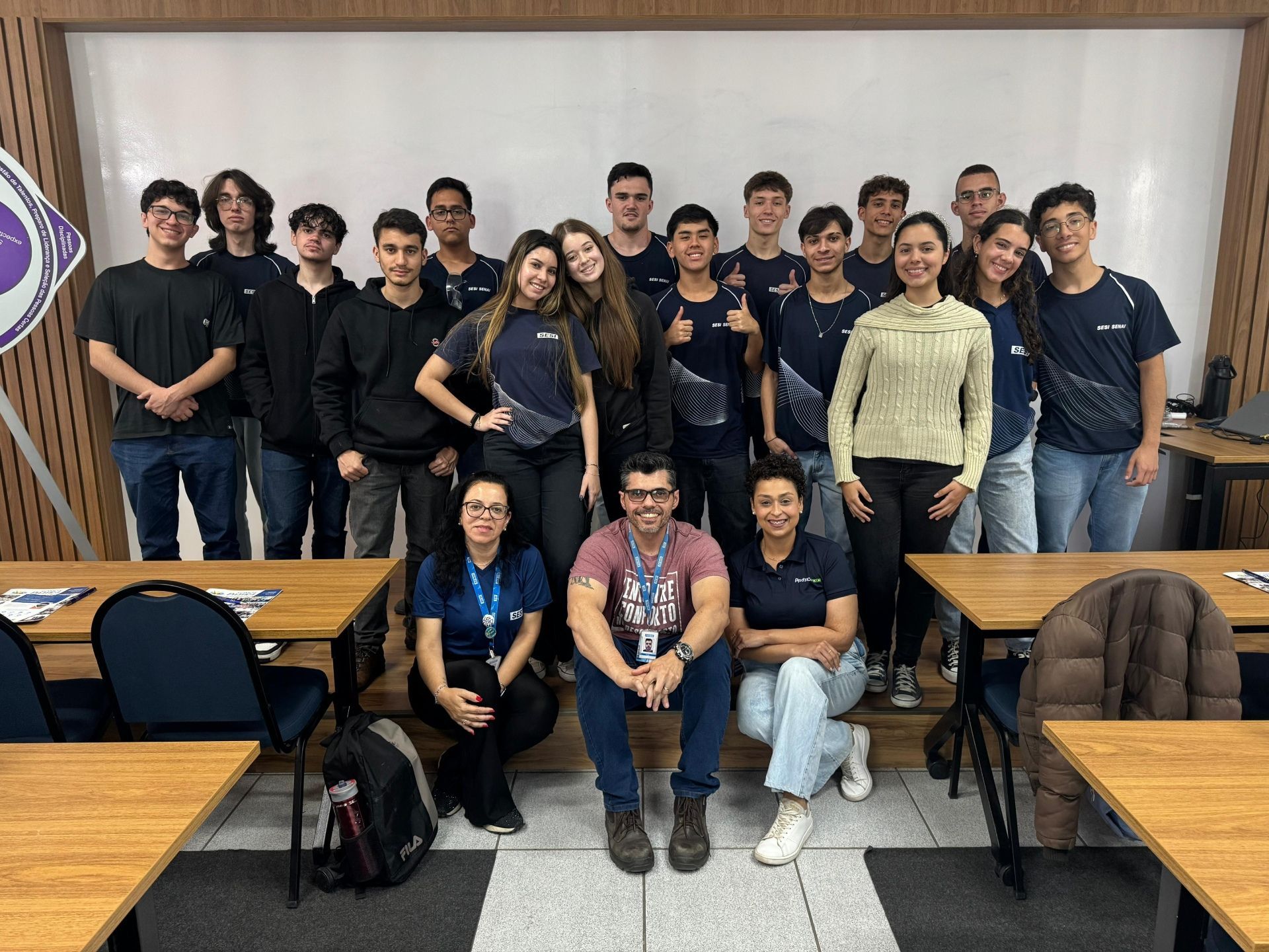 Foto de grupo: Alunos e instrutores em uma sala de aula, a maioria vestindo camisas azul-marinho.