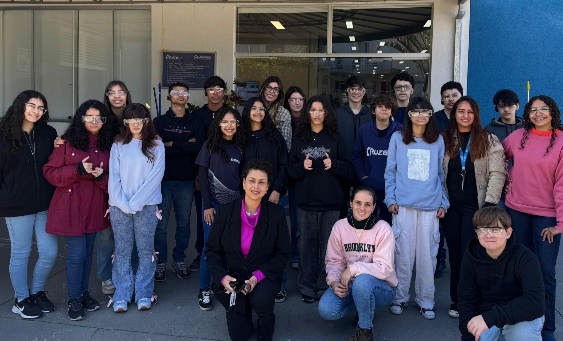 Um grupo de estudantes diversos posa com uma mulher ao ar livre. Eles sorriem para a câmera em um dia ensolarado.